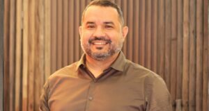 homem com barba grisalha olhando para frente e sorrindo usando camisa marrom claro
