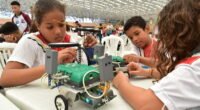 três crianças com um protótipo de robô sobre uma mesa