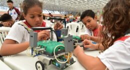 três crianças com um protótipo de robô sobre uma mesa