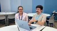 home de bigode sentado em frente a um notebook e com uma mulher ao lado