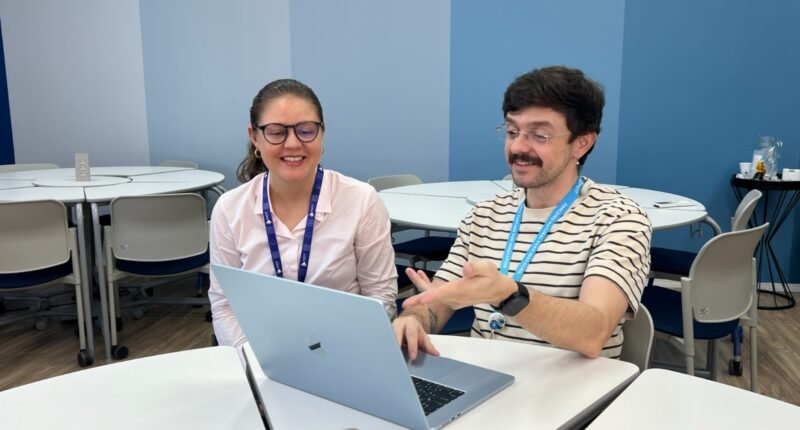 home de bigode sentado em frente a um notebook e com uma mulher ao lado