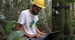 homem de capacete amarelo sentado no meio da floresta com um notebook no colo
